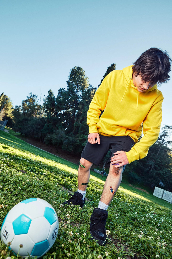 Teen in a yellow hoodie on grass beside a soccer ball, bent over checking their shin