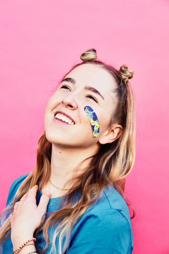 Teen in a blue t-shirt smiling against a pink background with a colorful World of Hurt bandage on their cheek