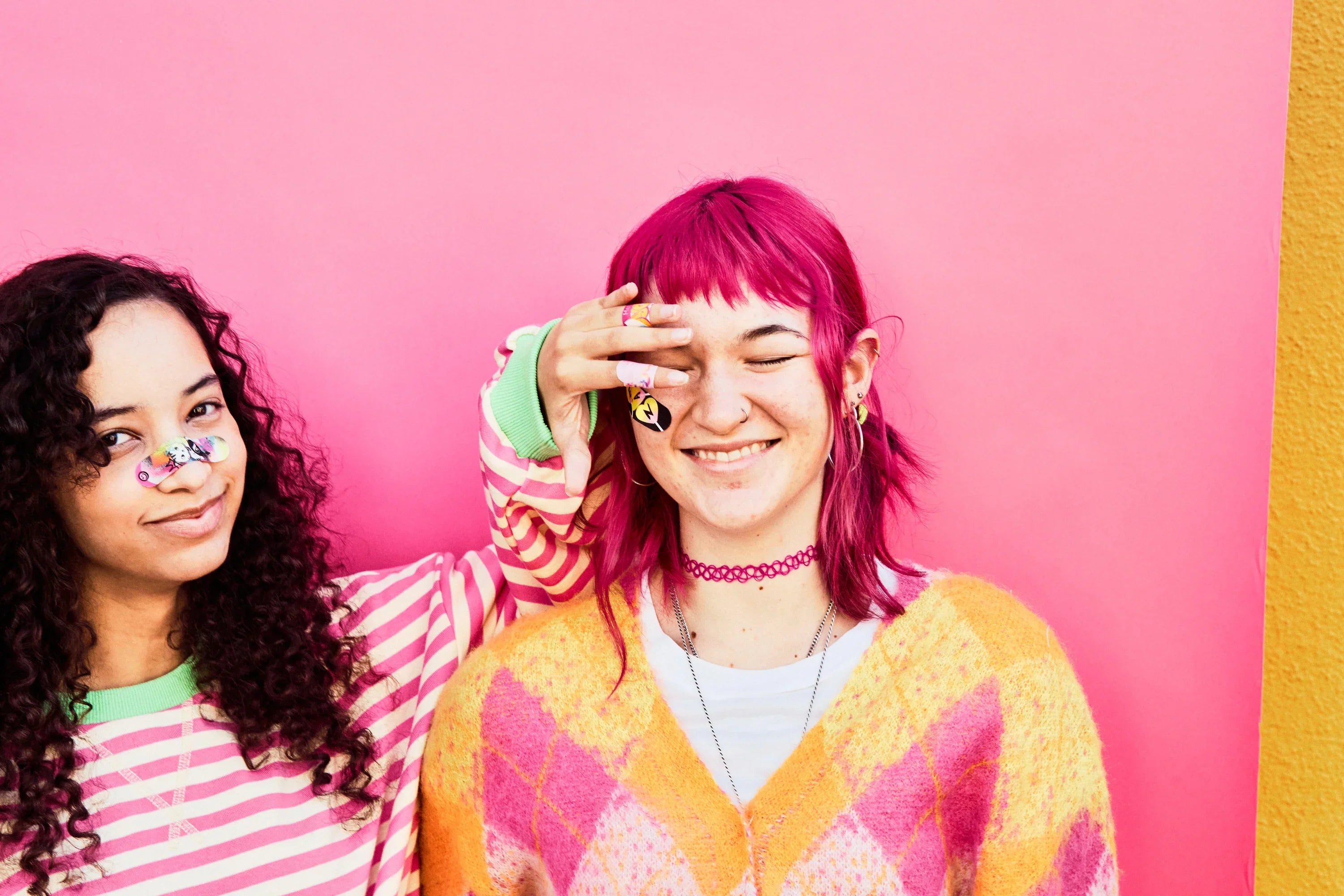 Two teens against a pink background, one wearing World of Hurt bandages on her cheek